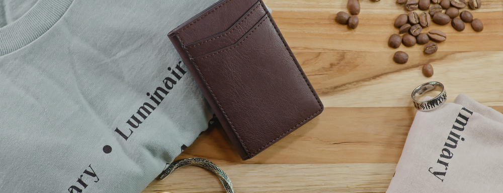 Flat lay shot of LuminaryLA products against a wood panel background with coffee beans. Products include a light blue hoodie, tan shirt, brown leather wallet, card holder, gray and silver bracelets, and silver ring.