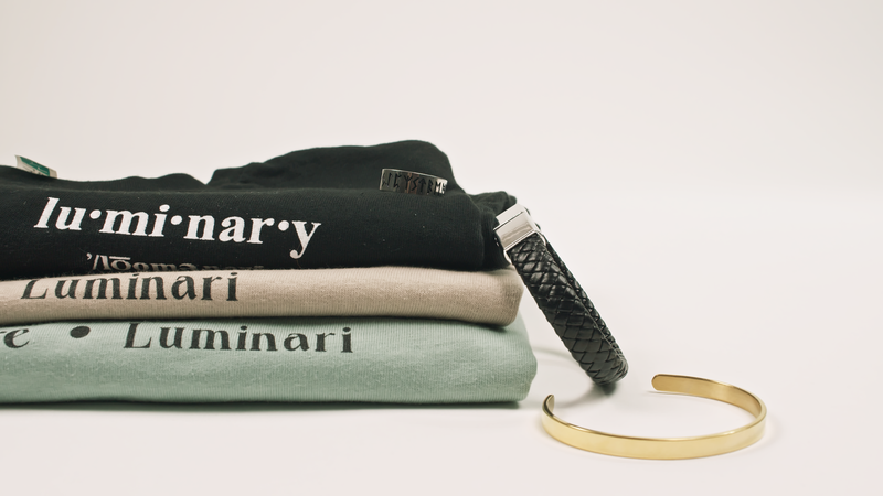 Product shot of neatly folded LuminaryLA shirts stacked, accompanied by a golden metal bracelet and a black leather bracelet, against a clean backdrop.