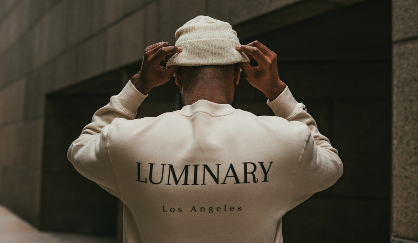 Rear view of model in tan LuminaryLA crewneck, adjusting beanie, with 'Luminary' and 'Los Angeles' in modern font on the back.