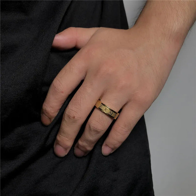 Close-up shot of a model's hand wearing a minimalist gold ring engraved with Roman numerals, resting against a plain black shirt.
